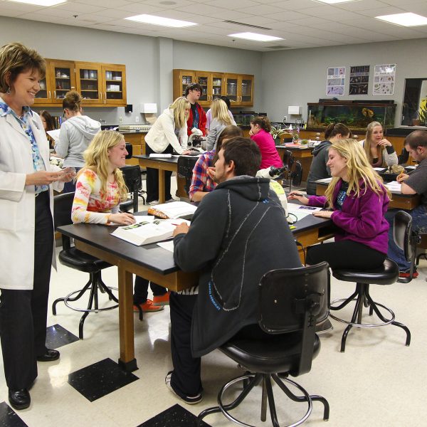 Instructor, in a lab coat, is speaking to her Life Science class.