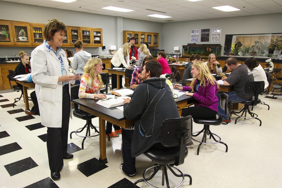 Instructor, in a lab coat, is speaking to her Life Science class.